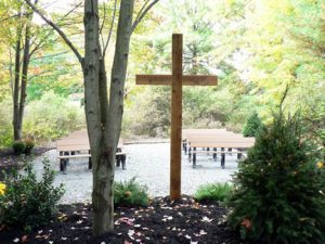 Wooden Cross in front of wooden pews surrounded by trees.